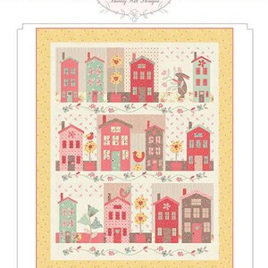 May include: A patchwork quilt featuring a whimsical village scene with colorful houses, flowers, and a bunny. The quilt has a yellow border and the words "Sunshine Lane" at the bottom. The Bunny Hill Designs logo is at the top.