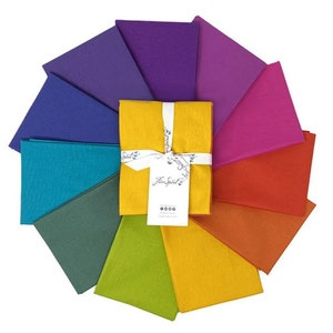 May include: A collection of colorful fabric squares arranged in a circular pattern. The squares are in shades of blue, green, yellow, orange, pink, and purple. A bundle of yellow fabric squares is tied with a white ribbon and a tag that reads "Fine Spirit Co."