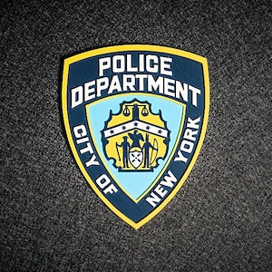 May include: A shield-shaped patch featuring the words "POLICE DEPARTMENT CITY OF NEW YORK" in white text. The patch has a blue border and a yellow outline. The center has a light blue background with a crest and two figures.