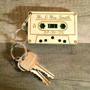 May include: A wooden cassette tape keychain engraved with "Mr. & Mrs. Smith" and the date "07-24-25". The keychain is attached to a silver keyring with several keys. The cassette tape design has a green center and black details.