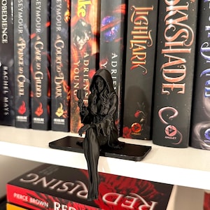May include: A matte black 3D printed figurine of a hooded figure sitting on a flat rectangular base. The figure wears a long robe with wide sleeves, has long wavy hair, and holds a small goblet in one hand while resting on a bookshelf.