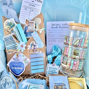 May include: A blue and white gift basket filled with book-themed items, including a reading list, a book review sheet, a sticker that says "I'd rather be reading", a mug with a bookshelf design, a necklace with a book charm, and a sticker that says "So many books".