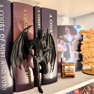 May include: A black figurine of a winged male figure, leaning against a row of books. The books have titles like "A Court of Mist and Fury" and "A Court of Thorns and Roses." A miniature wooden nightstand is also visible.