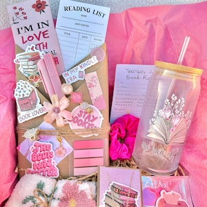 May include: A pink gift box filled with reading-themed items. Includes bookmarks, a reading list, pens, stickers, a scrunchie, a tumbler with a straw, and a pair of socks. The box is decorated with floral and book-related designs. Text includes "I'm in love with art", "Book Lover", and "The book was better".