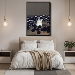 May include: A framed art print featuring a white cartoon character surrounded by many black, spiky creatures with large eyes. The artwork is set against a dark gray background. The print is displayed above a bed with white bedding and a tan throw.