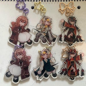 Könnte beinhalten: Eine Sammlung von Acryl-Anhängern mit Anime-Charakteren. Die Anhänger zeigen verschiedene Charaktere in detaillierten Outfits mit leuchtenden Farben und aufwendigen Designs. Jeder Anhänger hat einen Metallverschluss zur Befestigung.