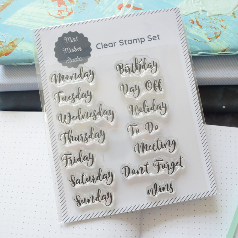 Set of clear stamps with calligraphy days of the week and other planner words like birthday and day off. The stamp set is leaning against a stack of notebooks.