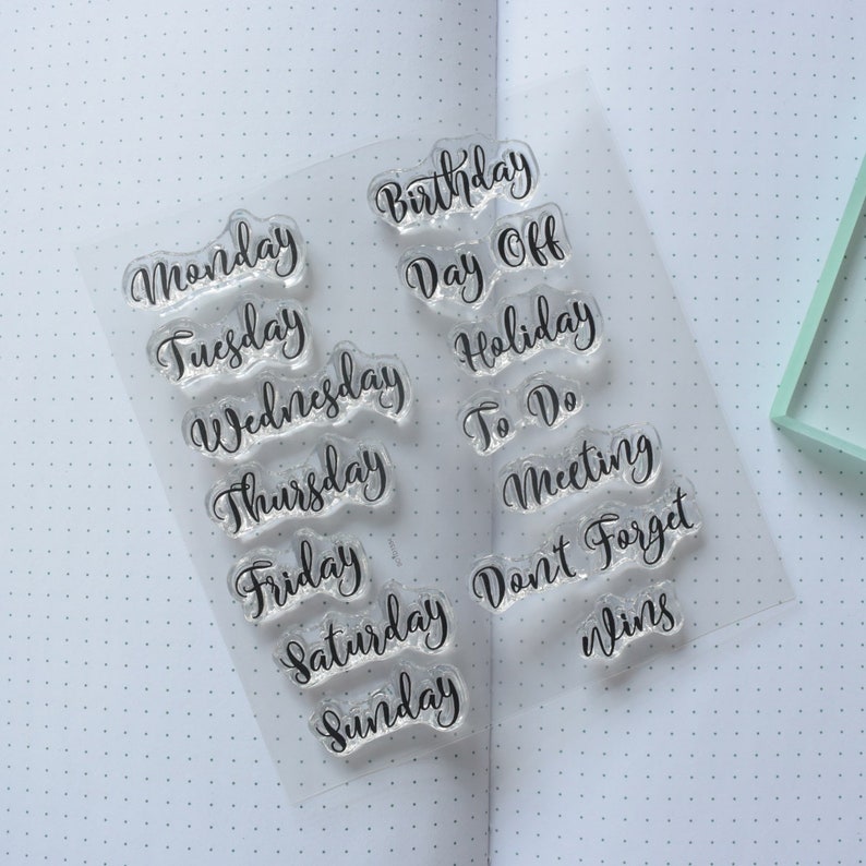 Set of clear stamps with calligraphy days of the week and other planner words like birthday and day off. The stamp set is sitting on an open notbook.