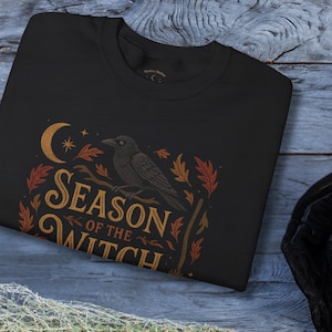 May include: Black sweatshirt with a 'Season of the Witch' design. The design features a crescent moon, stars, a raven perched on a branch, and autumn leaves in orange and gold. A black witch hat with a silver buckle and a broom are also visible.