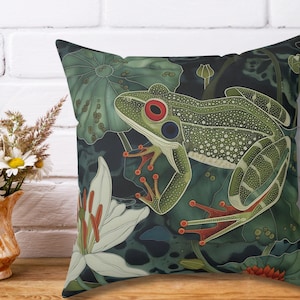 May include: A green frog with red eyes sits on a leafy green and black patterned throw pillow. The pillow has a white flower in the bottom left corner.