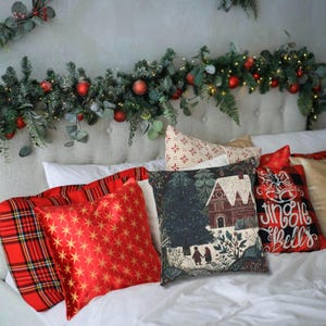 May include: A collection of Christmas-themed throw pillows in various designs and colors. Pillows include red plaid, gold starburst, and a pillow with a house and forest scene. A garland with red ornaments and greenery decorates the headboard.