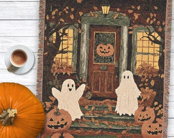 May include: A woven throw blanket featuring a Halloween scene with two white ghosts, multiple orange jack-o'-lanterns, and a house entrance with a pumpkin wreath on the door, all surrounded by dark trees and scattered autumn leaves.