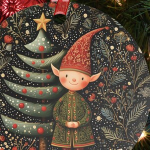May include: Round Christmas ornament featuring a whimsical elf with a red hat and green coat, standing in front of a stylized Christmas tree. The background is a dark, floral pattern with a star at the top. A red ribbon is attached for hanging.