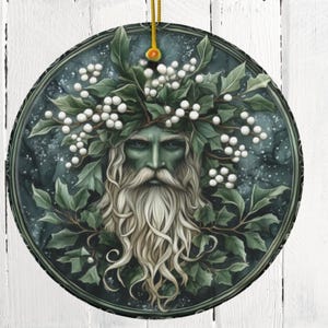 May include: A round, green and white Christmas ornament featuring a stylized image of the Green Man, a mythical figure associated with nature and the changing seasons. He is depicted with a beard and hair made of leaves and berries, and his face is framed by a wreath of holly.