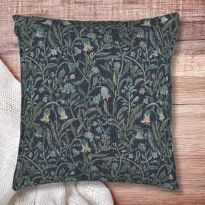 May include: Square decorative pillow with a dark blue background and a floral pattern in shades of teal, green, and beige. The pillow has a textured appearance and is placed on a wooden surface.