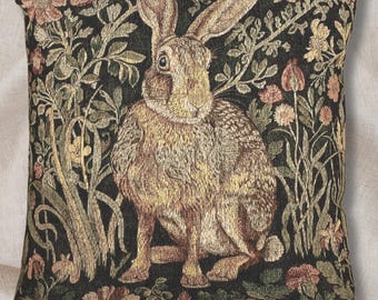 May include: Square decorative pillow featuring a detailed tapestry design. The pillow showcases a brown rabbit amidst a backdrop of flowers and foliage in shades of green, beige, and pink against a black background. The pillow has a soft, textured appearance.