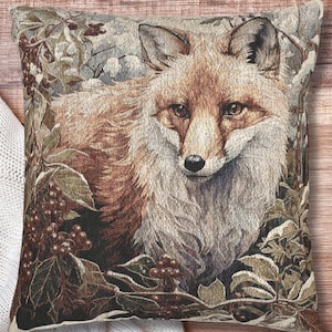 May include: Square decorative pillow featuring a detailed tapestry of a fox. The fox has reddish-brown fur and is surrounded by foliage in shades of green, brown, and beige. The pillow has a rustic, nature-inspired theme.