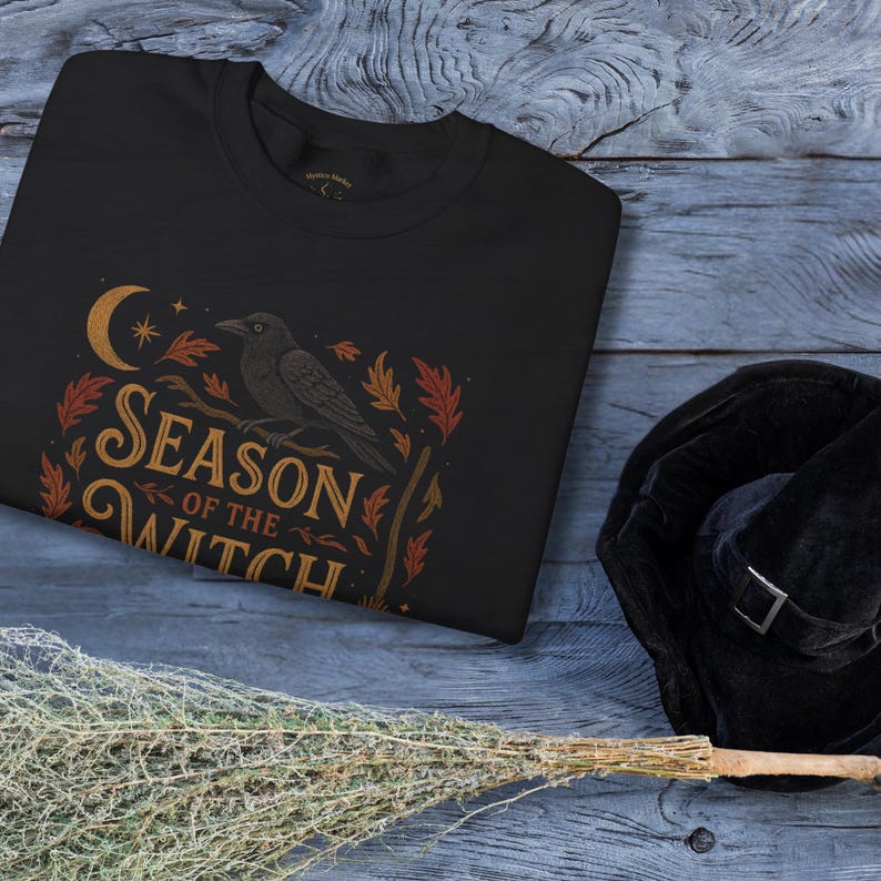 May include: Black sweatshirt with a 'Season of the Witch' design. The design features a crescent moon, stars, a raven perched on a branch, and autumn leaves in orange and gold. A black witch hat with a silver buckle and a broom are also visible.