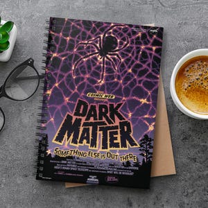 May include: A spiral-bound notebook with a cover design featuring the words "DARK MATTER" and the phrase "SOMETHING ELSE IS OUT THERE." The design includes a spiderweb pattern. The notebook is on a grey surface with a cup of coffee and glasses.