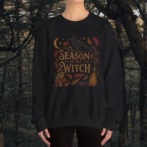 May include: Black sweatshirt with a graphic design featuring the text "Season of the Witch". The design includes a crescent moon, a crow, autumn leaves, a broom, and a mushroom with a frog.