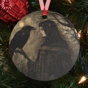 May include: Round Christmas ornament featuring a woman in a black hooded cloak with a raven perched on a branch. The artwork is in shades of gray and black, with a full moon in the background. A red ribbon is attached for hanging.