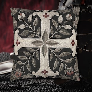 May include: Square decorative pillow with a dark background and a large, light-colored cross design. The cross is adorned with floral and leaf patterns in shades of gray and red. The pillow has a textured appearance.