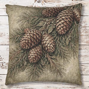 May include: Square decorative pillow featuring a detailed illustration of pine cones and needles. The pillow has a neutral beige background with a textured appearance. The pine cones are brown, and the needles are green.