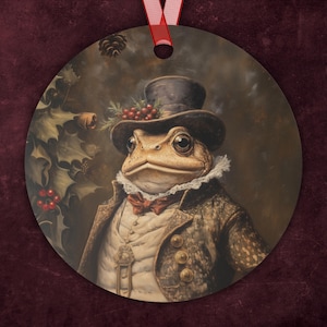 May include: Round Christmas ornament featuring a distinguished frog in a top hat, waistcoat, and jacket. The frog is adorned with a bow tie and a ruffled collar. Holly and berries decorate the hat. A red ribbon is attached for hanging.