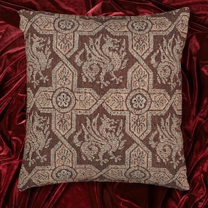 May include: Square decorative pillow with a detailed woven pattern in shades of brown and beige. The design features repeating geometric shapes, floral elements, and stylized mythical creatures. The pillow is placed on a red velvet surface.
