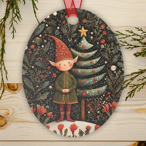 May include: Oval Christmas ornament featuring a whimsical elf in a green coat and red hat, standing next to a decorated Christmas tree. The background is a dark floral pattern with red accents, and a red ribbon is attached for hanging.