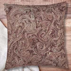 May include: Square decorative pillow with a swirling, marbled pattern in shades of brown, burgundy, and cream. The pillow has a textured appearance and is placed on a wooden surface next to a white knitted blanket. The pillow is a home decor item.