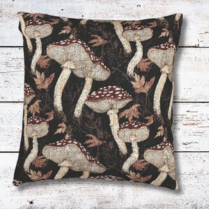 May include: Square decorative pillow with a repeating pattern of beige and burgundy mushrooms with white spots on a dark brown background. The pillow also features brown leaves. The pillow is placed on a white wooden surface.