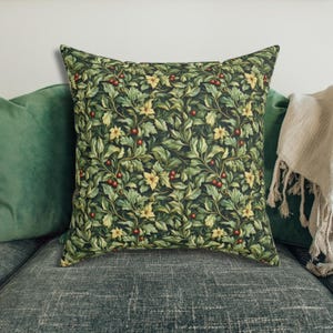 May include: Square decorative pillow with a botanical pattern. The pillow features a dark green background with a repeating design of green leaves, yellow flowers, and red berries. The pillow is placed on a dark gray couch with green pillows.