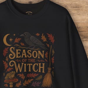 May include: Black sweatshirt with a graphic design featuring a crow perched on a branch, a crescent moon, and the words "SEASON OF THE WITCH." The design includes autumn leaves, a broom, and a toadstool.