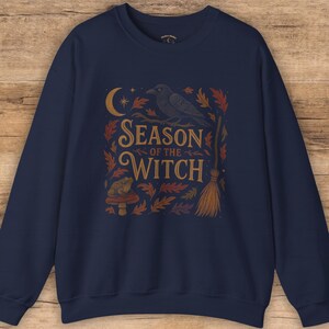 May include: Navy blue sweatshirt with a graphic design featuring a crow perched on a branch, a crescent moon, and the words "Season of the Witch." The design also includes a mushroom, leaves, and a broom.
