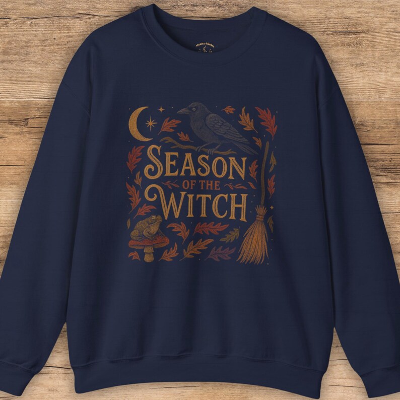 May include: Navy blue sweatshirt with a graphic design featuring a crow perched on a branch, a crescent moon, and the words "Season of the Witch." The design also includes a mushroom, leaves, and a broom.