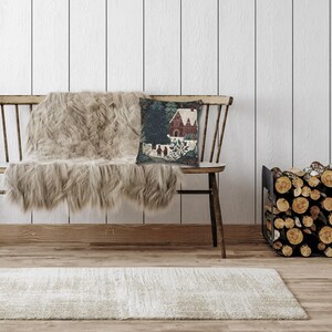 May include: A cozy interior scene featuring a wooden bench with a faux fur throw and a decorative pillow. The pillow displays a quaint house scene. A stack of firewood sits nearby, and a rug lies on the floor. The walls are white with vertical wood paneling.