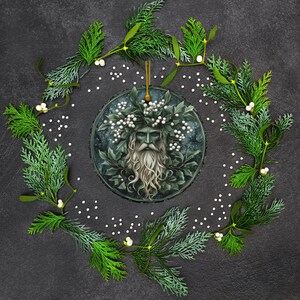 May include: A round, green and white Christmas ornament featuring a green man with a long white beard and a crown of leaves and white berries. The ornament is surrounded by a wreath of green leaves and white berries.