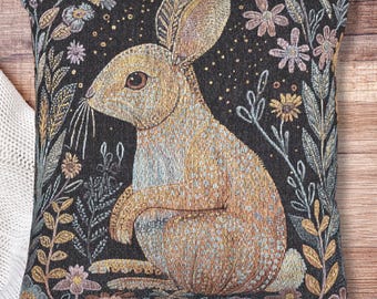 May include: Square decorative pillow featuring a detailed illustration of a rabbit surrounded by flowers and foliage. The rabbit is in shades of tan and brown, set against a dark background. The pillow has a woven texture and is a decorative home accent.