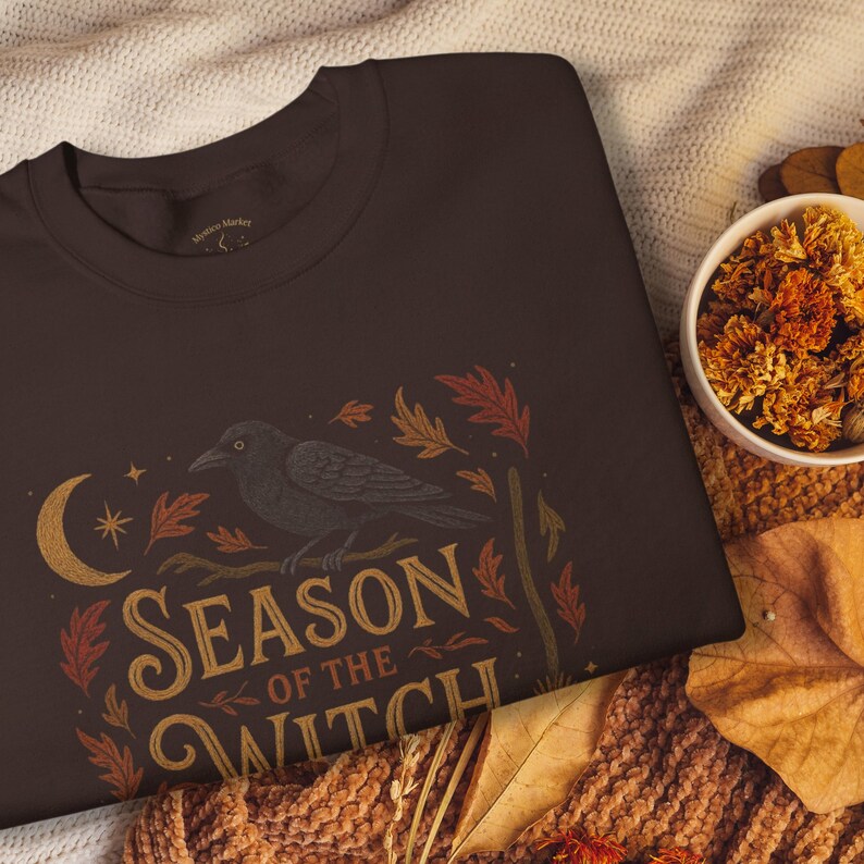 May include: A dark brown sweatshirt featuring a crow perched on a branch with the text "Season of the Witch." The design includes a crescent moon, stars, and autumn leaves in shades of gold, orange, and red.