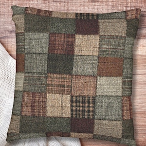 May include: Square decorative pillow with a patchwork design. The pillow features a variety of earth-toned squares in shades of green, brown, and beige. The squares have different textures and patterns, creating a rustic, cozy aesthetic.