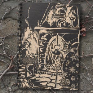 May include: A spiral-bound notebook with a black cover featuring a woodcut-style illustration. The artwork depicts a woman seated near a fireplace, with a witch-like figure in a cloak. The design is in shades of black and beige, with a rustic, vintage aesthetic.