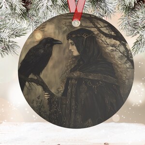 May include: Round metal ornament featuring a woman in a black hooded cloak with a raven perched on a branch. The artwork has a mystical theme, with a full moon in the background. A red ribbon is attached for hanging.