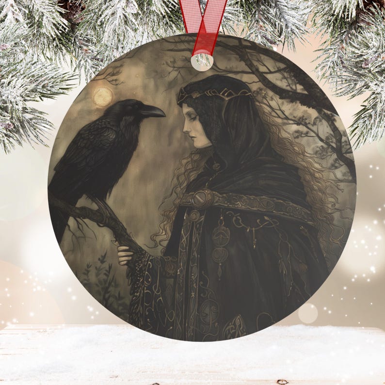 May include: Round metal ornament featuring a woman in a black hooded cloak with a raven perched on a branch. The artwork has a mystical theme, with a full moon in the background. A red ribbon is attached for hanging.