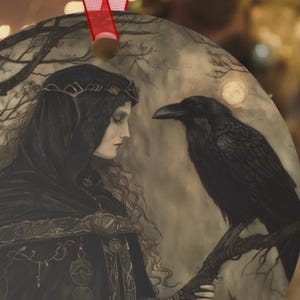 May include: Round ornament featuring a gothic illustration of a woman in a black hooded cloak and a raven perched on a branch. The artwork is in shades of gray and black, with a full moon in the background. A red ribbon is attached for hanging.