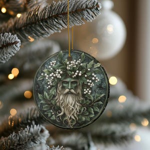 May include: A round ceramic ornament with a green man figure wearing a crown of holly and white berries. The background is a dark green and blue with a starry effect.