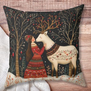 May include: Decorative square pillow featuring a whimsical illustration of a woman in a red dress embracing a white deer in a snowy forest. The deer has floral patterns and antlers. The background is dark with trees and red berries.