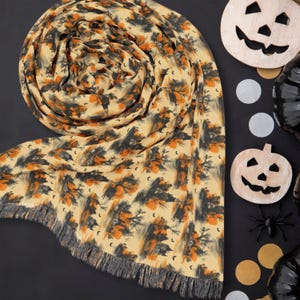 May include: A beige scarf featuring a repeating pattern of black haunted houses, bare trees, and orange accents, finished with a fringed edge. The fabric displays a detailed print of spooky silhouettes and small crescent moons across the surface.