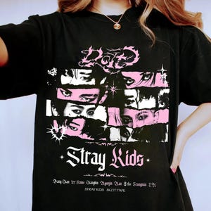 May include: Black t-shirt featuring a graphic design with the text "Stray Kids" in white and pink. The design includes stylized eyes and the names of the band members. The shirt is a casual, oversized style.