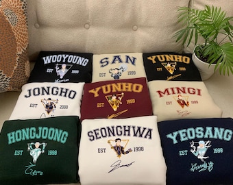 Puede incluir: Nueve sudaderas de color crema, negro y granate con diferentes nombres y fechas impresos en ellas. Los nombres incluyen Wooyoung, San, Yunho, Jongho, Mingi, Hongjoong, Seonghwa y Yeosang. Las fechas incluyen 1999 y 2000.